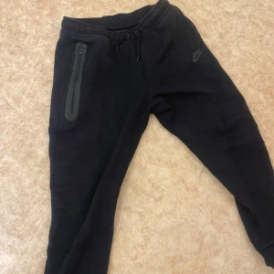 Svarta mjukisbyxor från Nike med dragkedja - Svarta mjukisbyxor från Nike med diskret logga på benet. Byxorna har dragsko i midjan och en praktisk ficka med dragkedja på sidan. Perfekta för chill eller träning, tillverkade i mjukt material för extra komfort.