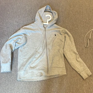 Grå hoodie med dragkedja från Polo Ralph Lauren - Säljer en klassisk grå hoodie från Polo Ralph Lauren med dragkedja framtill och huva med snörning. Tröjan har långa ärmar och en liten broderad logga på bröstet. Perfekt för en avslappnad stil.