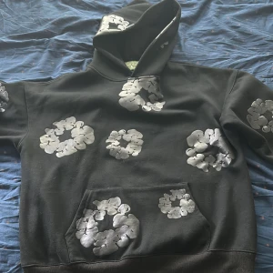 Svart hoodie med blommönster från Denim Tears - Säljer en svart hoodie från Denim Tears med unikt vitt blommönster tryckt över hela tröjan, inklusive huva och ärmar. Hoodien har klassisk känguruficka och ribbade muddar. Perfekt statement-plagg för dig som vill sticka ut. Använd cirka 2-3 gånger priset kan självklart diskuteras.