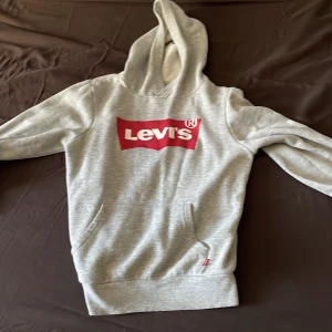 Grå hoodie från Levi's med röd logga - Säljer en klassisk grå hoodie från Levi's med stor röd logga på bröstet. Tröjan har huva, magficka och ribbade muddar vid ärmslut och nederkant. Perfekt för en avslappnad stil.