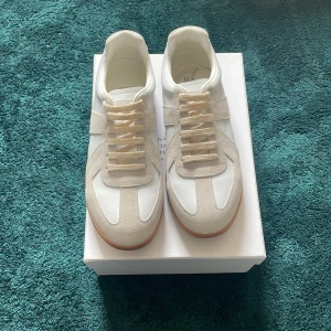 Priset går att diskutera - Säljer ett par stilrena Maison Margiela sneakers i vitt skinn och beige mocka med klassisk gummisula. Skorna har snörning och rund tå, perfekt för dig som gillar minimalistisk design och hög kvalitet.