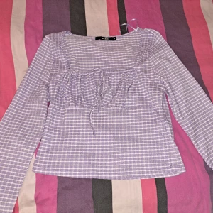 Ljuslila rutig blus med knyt - Säljer en söt ljuslila blus med vitt rutmönster och knyt framtill. Blusen har långa ärmar och en lätt croppad passform. Perfekt för dig som gillar romantisk och trendig stil.