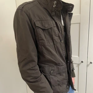 Mörkgrön utilityjacka från Massimo dutti  - Snygg mörkgrön utilityjacka från Massimo dutti med flera fickor framtill, hög krage med knappar och dragkedja. Jackan har en klassisk militärinspirerad look och är perfekt för lager-på-lager-stil under svalare dagar.