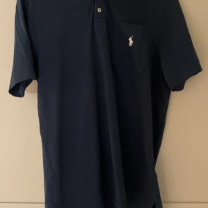 Mörkblå pikétröja från Ralph Lauren - Klassisk mörkblå pikétröja från Ralph Lauren med vit broderad logga på bröstet. Tröjan har korta ärmar, krage och knäppning framtill. Perfekt för en stilren och avslappnad look.