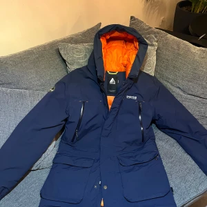 Blå dunjacka från 8848 Altitude - Säljer en blå dunjacka från 8848 Altitude med orange quiltat foder. Jackan har huva, dragkedja och flera fickor med dragkedjor. Perfekt för kalla dagar och har en sportig look med snygga detaljer. Den är som en helt ny jacka