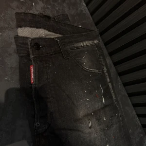 Dsquared2 jeans - Säljer ett par svarta jeansbyxor från Dsquared2 med tydliga slitningar och vita samt röda färgstänk på framsidan. Byxorna har klassisk femficksdesign, knappgylf och rakt snitt. Perfekta för dig som gillar en edgy och trendig stil.