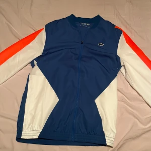  Lacoste dress - Säljer en snygg blå tränings dress från Lacoste Sport med vita och orange detaljer på ärmarna. Jackan har dragkedja framtill, lång ärm och klassisk Lacoste-logga på bröstet. Perfekt för sportiga tillfällen eller som streetwear. Köpt i Lacoste butiken i Kungsbacka 