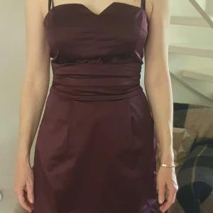 Lila kort klänning från Lindex - Säljer en lila kort klänning från Lindex med smala axelband och markerad midja. Klänningen har en hjärtformad urringning och är tillverkad i ett glansigt material som ger en elegant känsla.