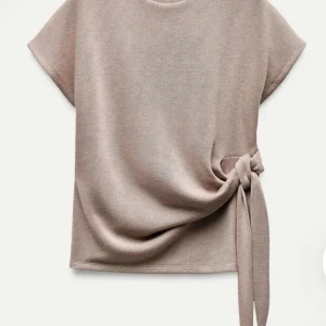 Vit topp med knyt från Zara - Säljer en stilren vit topp från Zara med korta ärmar och snygg knytdetalj i sidan. Toppen har en rund halsringning och är tillverkad i ett mjukt material som känns skönt mot huden. Perfekt för en enkel men trendig look.