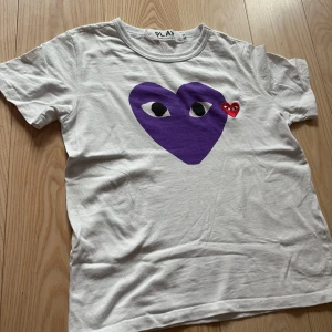 Vit t-shirt med lila hjärta från Comme des Garçons PLAY - Vit t-shirt från Comme des Garçons PLAY med stort lila hjärta och ögon på bröstet samt ett mindre rött hjärta bredvid. Klassisk rund halsringning och korta ärmar. Perfekt statement-plagg för dig som gillar unika prints. Storlek S