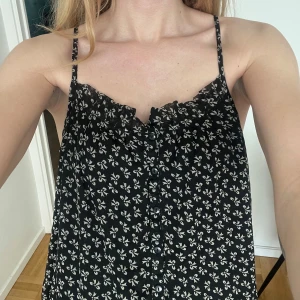 Svart linne med vita detaljer från VILA - Säljer en svart linne från VILA. Linnet har vitt rosettmönster och smala axelband. Det har knappar framtill och en lätt, luftig passform – samt diskret volangdetalj vid urringningen. Aldrig använt, så jättefint skick. 