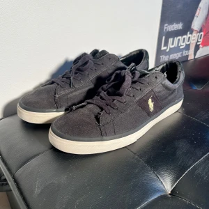 Svarta sneakers från Polo Ralph Lauren - Svarta sneakers från Polo Ralph Lauren med vit sula och klassisk gul broderad logga på sidan. Skorna har snörning och är tillverkade i textilmaterial. Perfekta för en stilren och avslappnad look. Storlek 44.5 