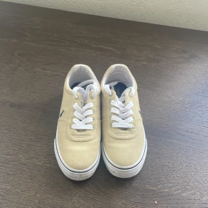 Beige sneakers från Polo Ralph Lauren - Säljer ett par beige sneakers från Polo Ralph Lauren med vit sula och vita skosnören. Skorna har en broderad mörkblå logga på sidan och texten POLO bak på hälen. Ovandelen är i textil och de har en klassisk, rund tå.