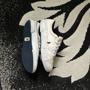 Vita sneakers från Polo Ralph Lauren - Säljer ett par vita sneakers från Polo Ralph Lauren med klassisk låg profil och snörning. Skorna har en mörkblå sula och den ikoniska Polo-loggan broderad på sidan. Perfekta för en clean och stilren look.