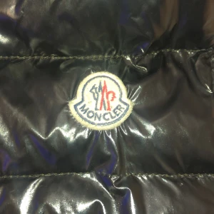 Svart dunväst från Moncler gui, storlek S - Snygg svart dunväst från Moncler med glansig finish och hög krage. Västen har dragkedja framtill, två fickor med dragkedja och Moncler-logga på bröstet. Perfekt för lager-på-lager och stilren look.