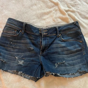 Mörkblå jeansshorts med fransar - Säljer ett par mörkblå jeansshorts med slitna detaljer och fransig kant nertill. Shortsen har klassisk femficksdesign, dubbla knappar i midjan och snygga slitningar både fram och bak. Perfekta för sommardagar!