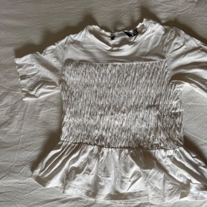 Vit topp med smock och volang - Säljer en söt vit topp med smockad front och rygg samt volang nertill. Toppen har rund halsringning och korta ärmar. Perfekt för en fräsch och somrig look!