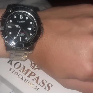 Svart dykarklocka från Kompass Stockholm - Stilren herrklocka från Kompass Stockholm med svart urtavla, datumvisning och tydliga index. Boett och länk i silverfärgat metall, vattentät till 100 meter och quartz-urverk. Perfekt accessoar för dig som gillar klassisk design. Låda finns 