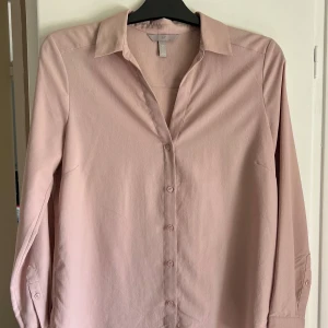 Ljusrosa skjorta från H&M - Säljer en stilren ljusrosa skjorta från H&M med klassisk krage och knäppning framtill. Skjortan har långa ärmar och är tillverkad i ett mjukt, lätt material som passar perfekt till många olika outfits. Aldrig använd 