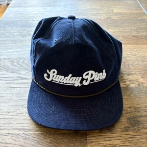 Mörkblå keps från Adidas med Sunday Pins-brodyr - Mörkblå keps i manchester med vit broderad text 'Sunday Pins' framtill och justerbart läderband bak. Klassisk design med repdetalj ovanför skärmen. Perfekt för en avslappnad stil.