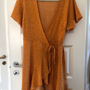 Prickig omlottklänning i orange  - Superfin orange omlottklänning med vita prickar och knyt i midjan. Klänningen har korta ärmar, v-ringning och volangdetalj nertill. Perfekt till sommaren! 🧡 Vet inte exakt vilken storlek det är på den, men skulle säga xs/s 🧡
