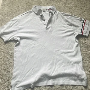 Vit piké från Moncler med logga - Säljer en klassisk vit pikétröja från Moncler i storlek L. Tröjan har korta ärmar, knappar vid halsen och Moncler-logga på ärmen. Ränder i rött, vitt och blått på ärmslutet ger en snygg detalj. Perfekt för en stilren look.