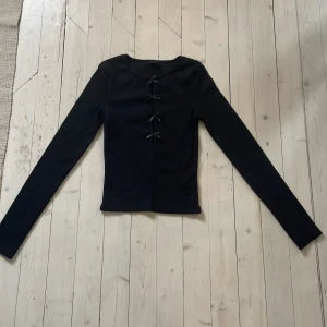 Svart långärmad topp med rosetter från Gina Tricot - Säljer en svart långärmad topp från Gina Tricot med dekorativa rosetter framtill. Toppen har en tight passform och är perfekt för att ge en stilren look med en söt detalj framtill.