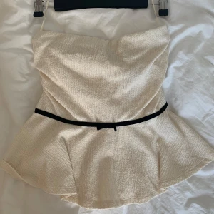 Beige axelbandslös topp från Holly & Whyte - Säljer en superfin beige axelbandslös topp från Holly & Whyte by Lindex. Den har ett svart band i midjan med en gullig rosett på. Oanvänd. 