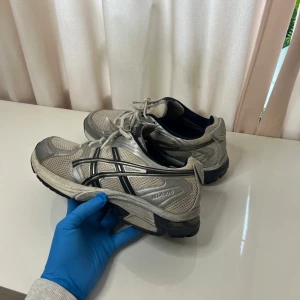 Asics - Säljer ett par gråa Asics sneakers med svarta och silvriga detaljer. Skorna har meshpaneler för extra ventilation, snörning framtill och en robust sula. Perfekta för träning eller vardagsbruk.