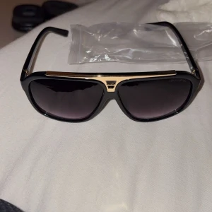 Svarta solglasögon från Louis Vuitton - Snygga svarta solglasögon från Louis Vuitton med guldiga detaljer upptill. Modellen har en klassisk pilotform och mörka glas. Perfekta för att lyfta din stil under soliga dagar. De är sällsynta finns knappt nånstans.