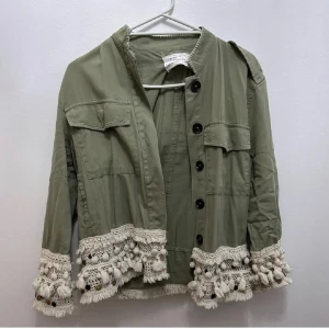 Grön jacka med virkade detaljer och tofsar - Pris kan diskuteras❣️ Snygg grön jacka med dekorativa virkade spetsdetaljer och tofsar längs ärmslut och nederkant. Jackan har knappar framtill, två bröstfickor och lång ärm. Perfekt för dig som vill sticka ut med en unik stil.