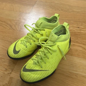 Gula inomhusfotbollsskor från Nike - Säljer ett par gula inomhusfotbollsskor från Nike med snörning och en högre ankelkrage för extra stöd. Skorna har en svart Nike-logga och är designade för att ge bra grepp på planen. 