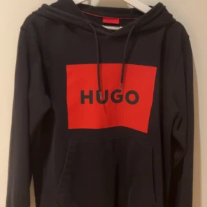 Svart hoodie från Hugo Boss - Snygg svart hoodie från Hugo Boss med röd logga på bröstet. Tröjan har en klassisk huva med dragsko och långa ärmar. Perfekt för en avslappnad stil.