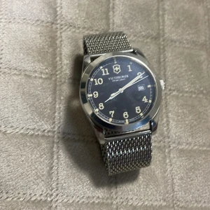 Silverfärgad klocka från Victorinox - Stilren klocka från Victorinox med en svart urtavla och tydliga siffror. Klockan har ett datumfönster och ett silverfärgat mesh-armband som ger en modern look. Perfekt för den som gillar en klassisk men ändå trendig stil.