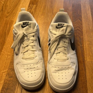 Vita Nike sneakers med svart swoosh - Snygga vita Nike sneakers med svart swoosh och snörning. Perfekta för en stilren look. Skorna har en klassisk design med Nike-logga på hälen och tungan.