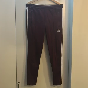 Vinröda träningsbyxor från Adidas - Snygga vinröda träningsbyxor från Adidas med vita ränder längs sidorna. Byxorna har dragkedja vid fickorna och Adidas-logga på ena benet. Perfekta för en sportig look.