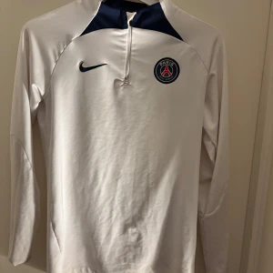 Vit långärmad tröja från Nike med PSG-logga - Säljer en vit långärmad tröja från Nike med PSG-logga på bröstet. Tröjan har en dragkedja vid halsen och är tillverkad i Dri-FIT-material för optimal komfort. Perfekt för fotbollsfans! ⚽