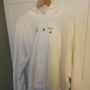 Vit hoodie från Off-White - Säljer en vit hoodie från Off-White med tryck på framsidan och ett blått grafiskt mönster på baksidan. Hoodien har en oversize passform med långa ärmar och en bekväm huva. Perfekt för en stilren och modern look. Orignalpåse och taggar följer med