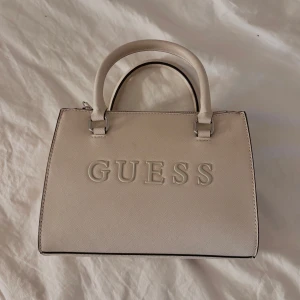 Beige väska från Guess - Säljer en beige handväska från Guess. Köpt för två år sedan på Guess butiken i Barkarby. Säljer för att jag helt enkelt slutat använda den. Köpt för 1500kr. Har ytterst 2 små fläckar på sig. 