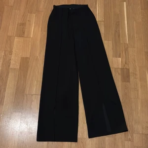Svarta byxor från Gina Tricot - Snygga svarta byxor från Gina Tricot med hög midja och vida ben. Byxorna har en stilren design med slitsdetaljer vid fållen. Perfekta för en chic look.