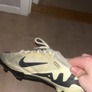 Beige fotbollsskor från Nike elite air zoom - Säljer ett par beige fotbollsskor från Nike med svarta detaljer och snörning. Skorna är elite och nypris cirka 3100 kronor men säljs för enbart 1200 för dom är använda en halv säsong och jätte bra skor att lira med om man spelar fotboll 