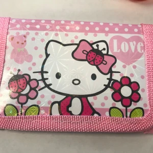 Rosa Hallo Kitty plånbok - Söt rosa plånbok med Hello Kitty-motiv. Plånboken har ett lekfullt design med prickar och hjärtan, samt bilder av Hello Kitty med olika teman. Perfekt för den som älskar färgglada och charmiga accessoarer. 55kr/st , helt ny 