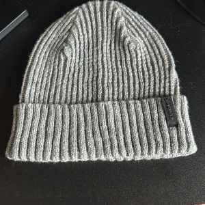 Grå ribbad mössa från Acne Studios - Snygg grå ribbad mössa från Acne Studios. Perfekt för kyliga dagar med sin stilrena design och mjuka material. Mössan har en uppvikt kant och en diskret etikett på sidan.