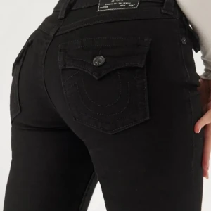 True Religion Svarta Joey Low Rise Flare - Svarta jeans från True Religion med låg midja och flare-stil. Byxorna har dekorativa sömmar och knappar på bakfickorna. Perfekta för en trendig look. Lite slitna ner till, sitter som w26