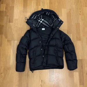 Burberry jacka  - Säljer en snygg svart dunjacka från Burberry  med klassiskt rutigt mönster i fodret. Jackan har en praktisk huva och stängs med både dragkedja och knappar. Man kan också ta av armarna på den så att man kan ha den som en väst. Perfekt för alla väder! 
