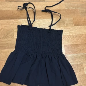 Svart axelbandslös topp - Snygg svart axelbandslös topp med smockdetaljer och knytband vid axlarna. Perfekt för en stilren look. Den har en luftig och bekväm passform.