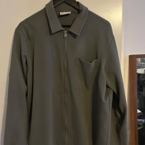 Grå overshirt med dragkedja - Snygg grå overshirt med dragkedja framtill och en bröstficka. Perfekt för lager-på-lager-stil. Den har en klassisk krage och långa ärmar.