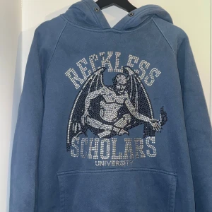 Blå reckless scholars - Säljer en blå hoodie med ett unikt tryck av en demon och texten 'Reckless Scholars University'. Hoodien har en klassisk design.