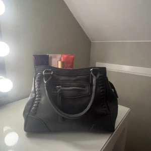 Svart handväska med dragkedja - Snygg svart handväska i skinn med dragkedja framtill. Väskan har två handtag och dekorativa sömmar längs sidorna. Perfekt för en stil ren look