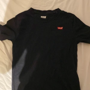 Svart t-shirt från Levi's - Snygg svart t-shirt från Levi's med klassisk röd logga på bröstet. Perfekt för en stilren look. Tillverkad i mjukt material för bekväm passform. 